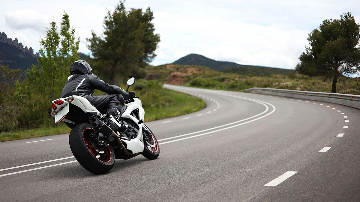Motorcyclist in black gear riding a white sportbike on a winding rural road surrounded by trees and hills under a cloudy sky.