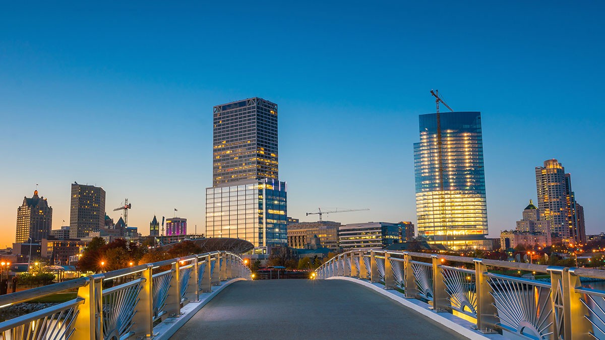 A city skyline at dusk with lit-up skyscrapers. The scene is viewed from a modern pedestrian bridge under a clear, deep blue sky.