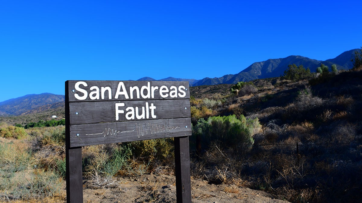 Sign reading "San Andreas Fault" with a mountainous landscape in the background under a clear blue sky.