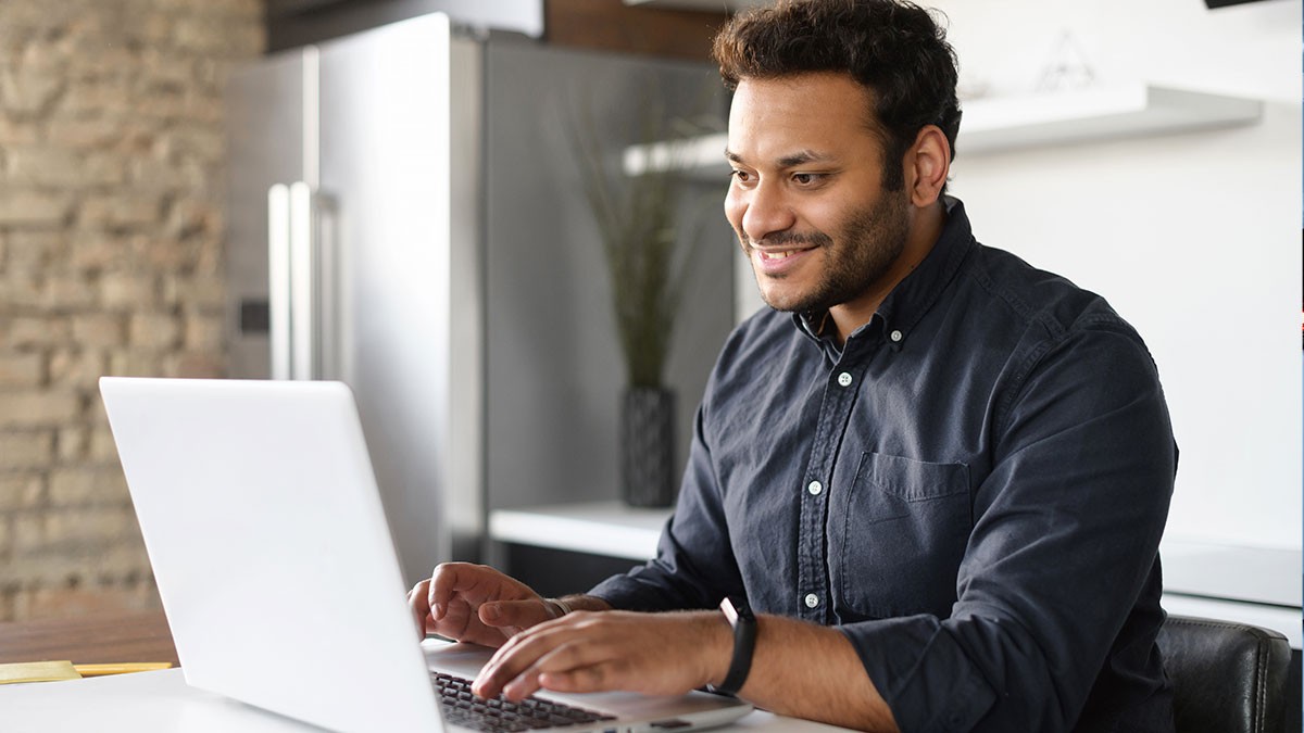Man smiling while typing on a laptop at a table in a modern kitchen setting.