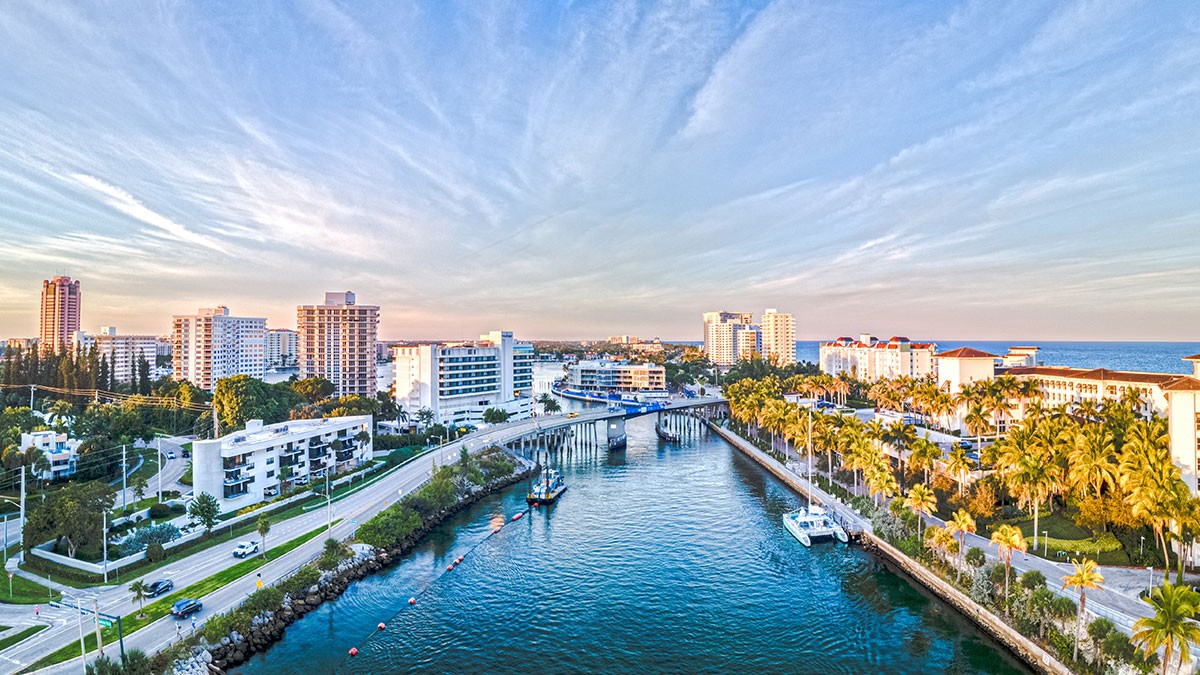 Aerial view of a coastal city with a bridge over a waterway, surrounded by buildings, palm trees, and a clear sky at sunset.