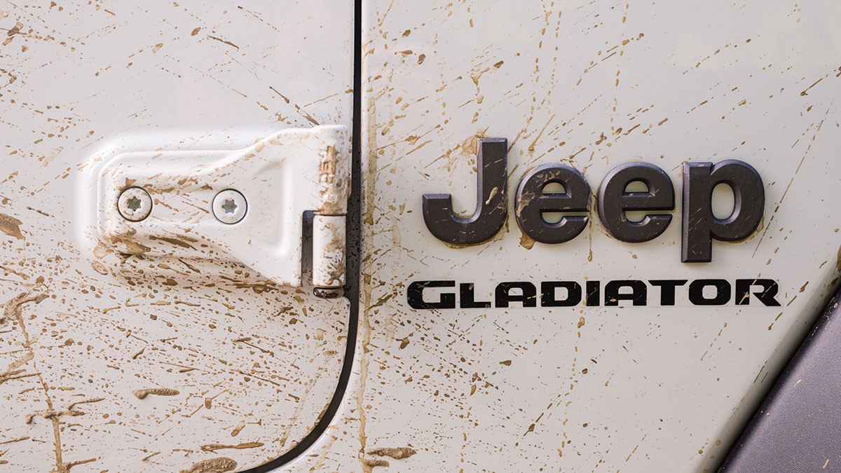 Close-up of a white Jeep Gladiator door with mud splatters, highlighting the Jeep logo and hinge.