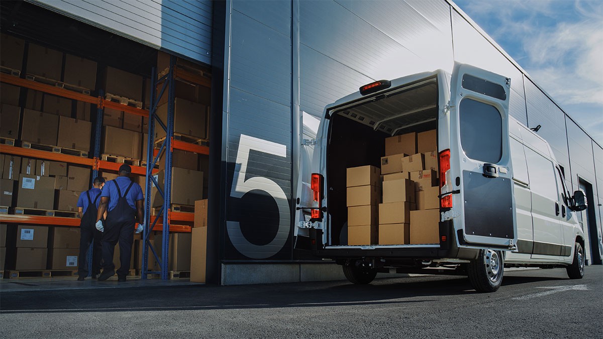 A van with open rear doors loaded with boxes is parked outside a warehouse, where two workers are stacking boxes on shelves.