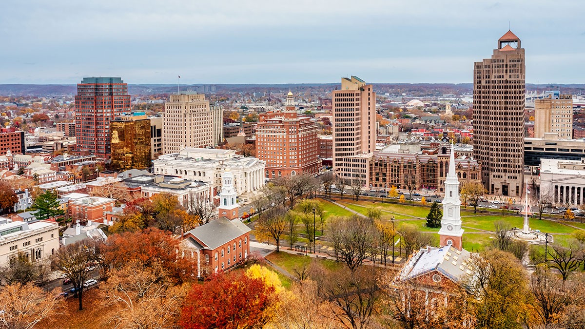Aerial view of a cityscape with historic buildings, a park with autumn trees, and modern skyscrapers under a cloudy sky.