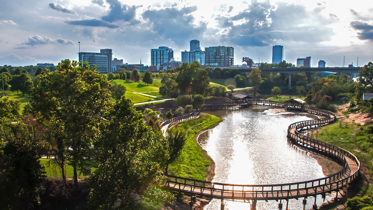 Scenic view of a winding river with a wooden boardwalk, lush greenery, and a city skyline under a cloudy sky.