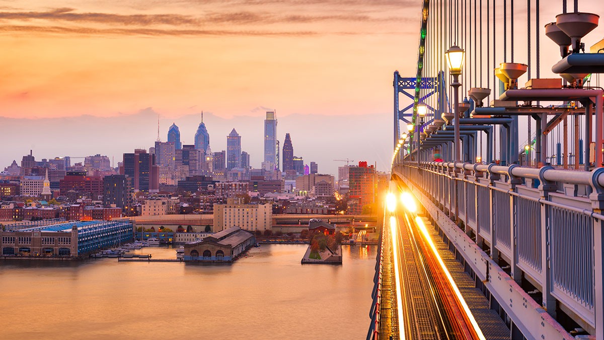 A city skyline at sunset with a bridge in the foreground, illuminated by streetlights, and a river reflecting the vibrant sky.