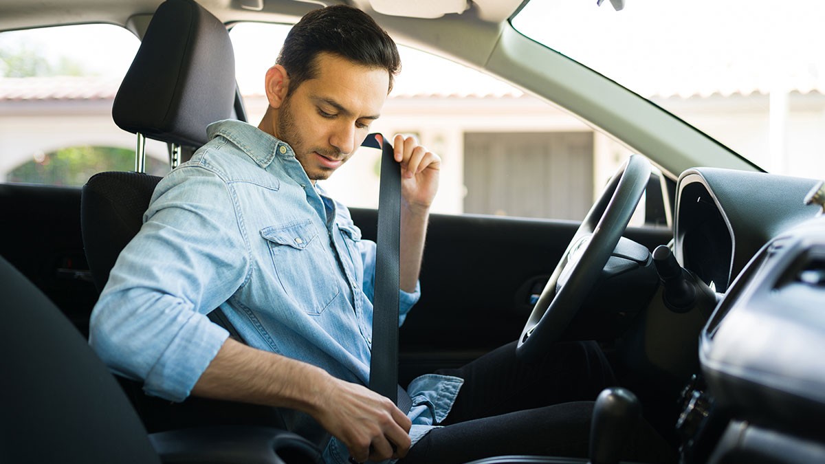 A man in a denim shirt fastens his seatbelt while sitting in the driver's seat of a car.