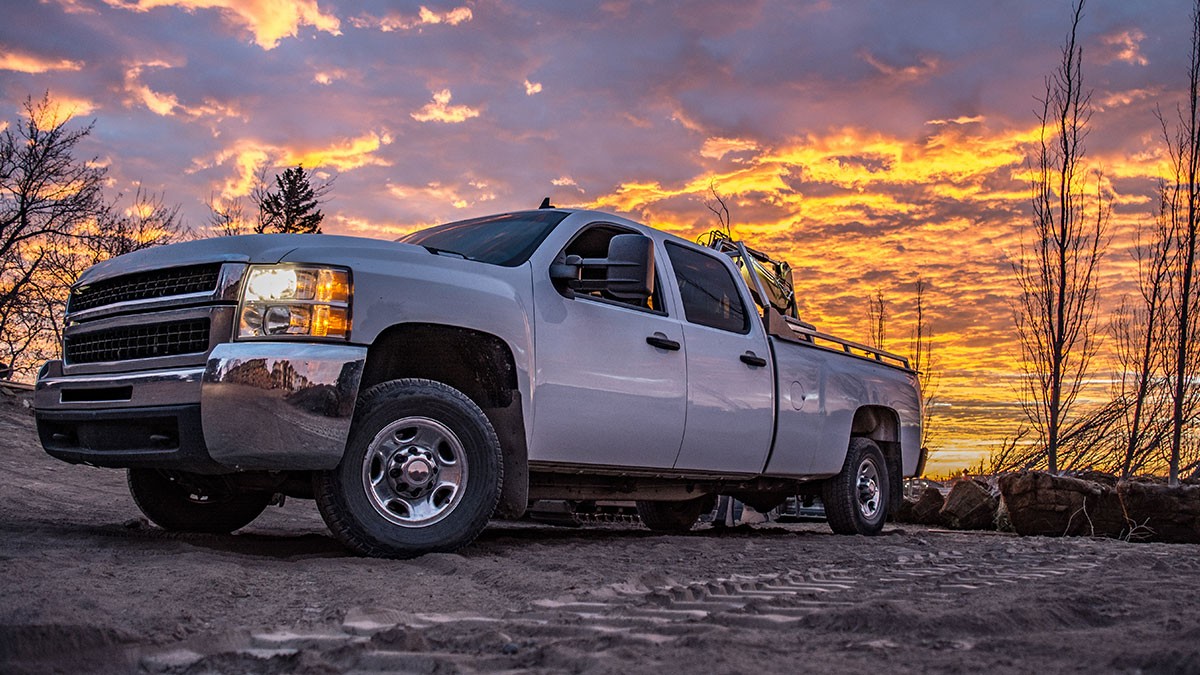White pickup truck parked on sandy ground, silhouetted against a vibrant sunset sky with dramatic clouds.