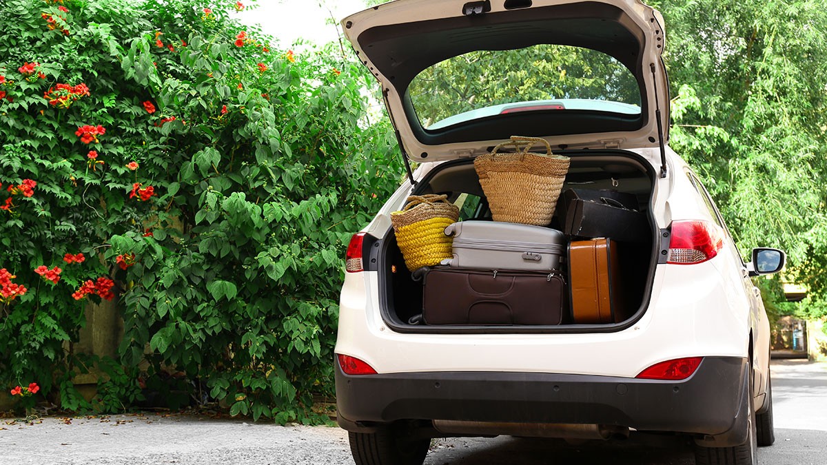 Open trunk of a white SUV packed with suitcases and woven baskets, parked near lush greenery with red flowers.