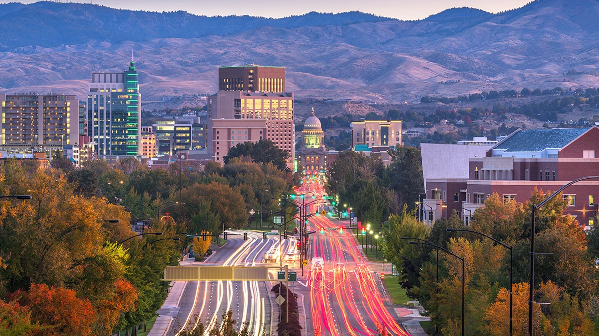 A cityscape at dusk with blurred car lights, modern buildings, and mountains in the background under a soft, colorful sky.