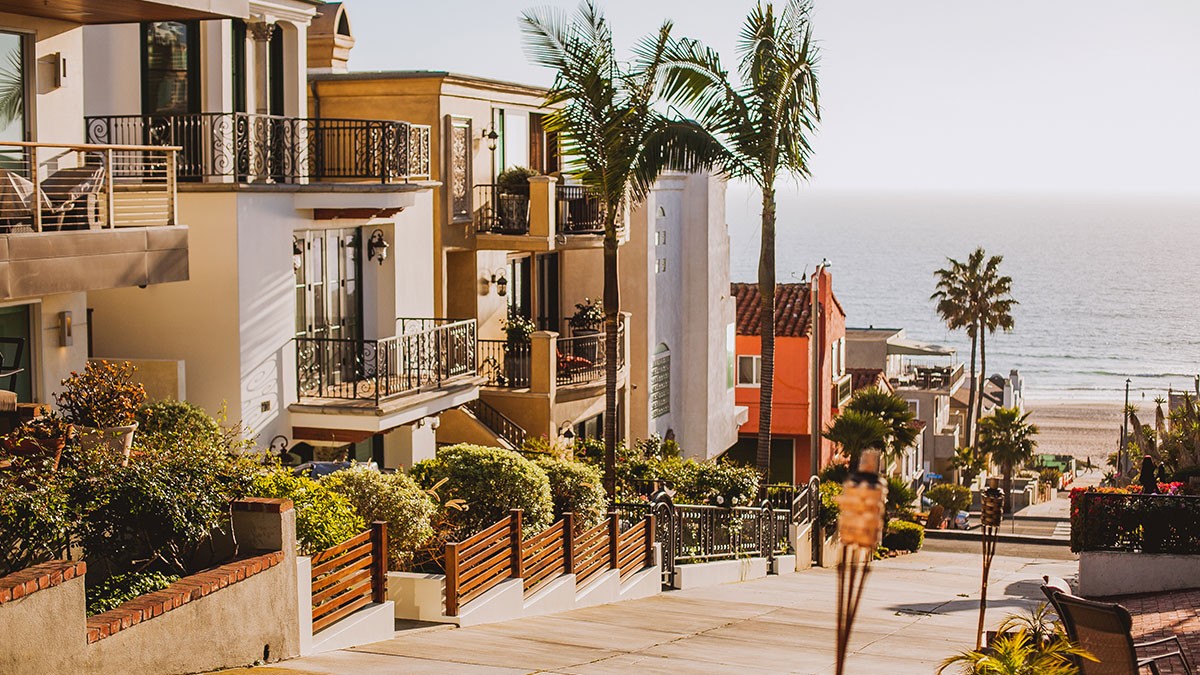 Coastal street scene with modern houses, lush greenery, and a view of the ocean in the background under a clear sky.