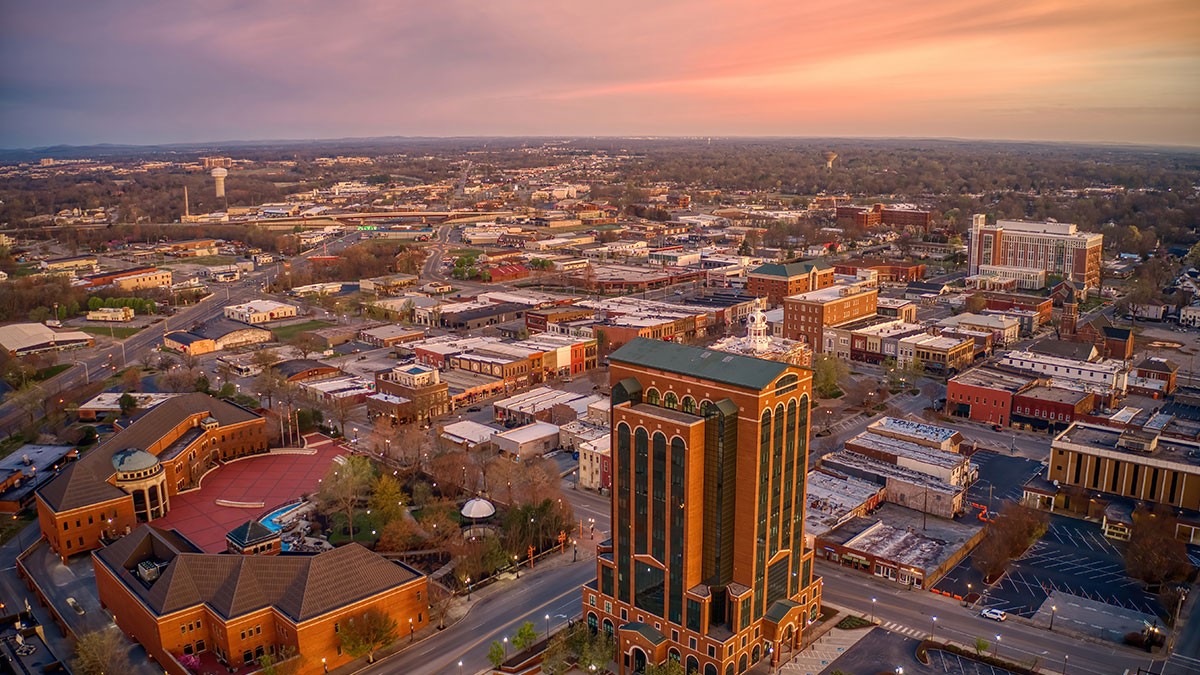 Aerial view of a cityscape at sunset, featuring a prominent tall building and surrounding urban architecture under a colorful sky.