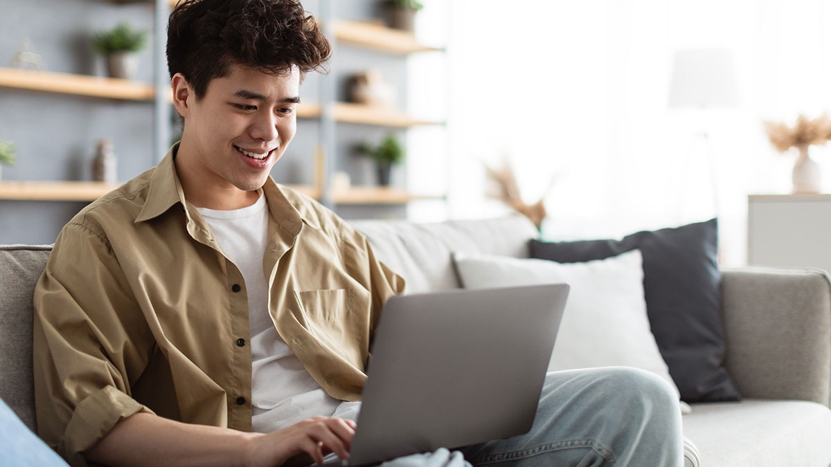 Smiling person sitting on a couch using a laptop in a cozy living room with shelves and soft lighting.