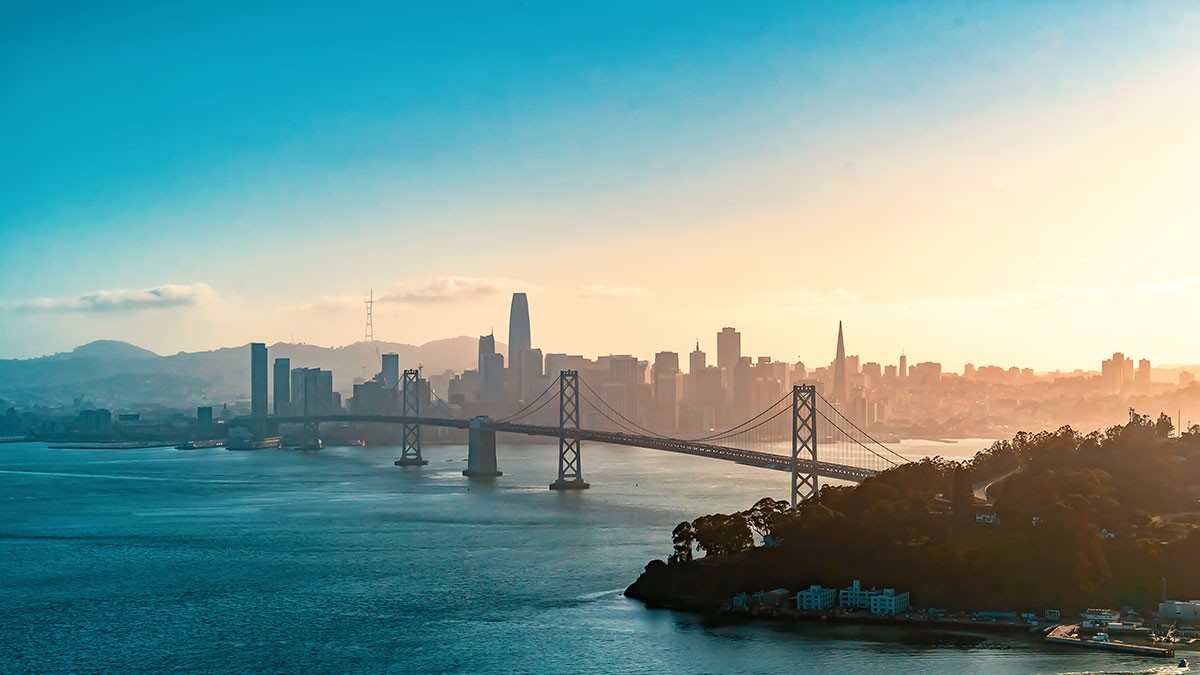 Aerial view of a city skyline at sunset with a prominent suspension bridge spanning across a bay, under a clear blue sky.