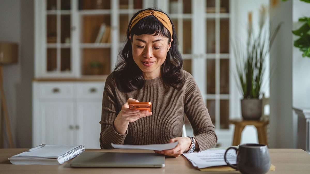 Woman sitting at a desk, smiling while looking at her phone. She's surrounded by a laptop, notebooks, papers, and a mug.