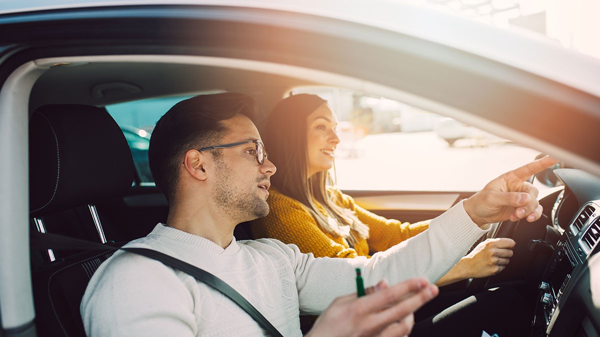 A man and woman are sitting in a car, with the woman driving and the man pointing ahead. Sunlight streams through the window.