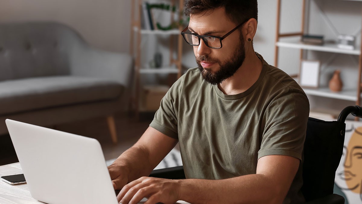 A man with a beard and glasses uses a laptop at a desk in a cozy room with a gray sofa and bookshelves.