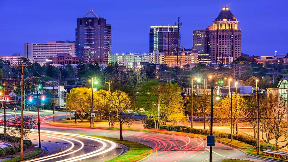 Cityscape at dusk with illuminated buildings, a winding road with light trails, and trees in the foreground under a deep blue sky.