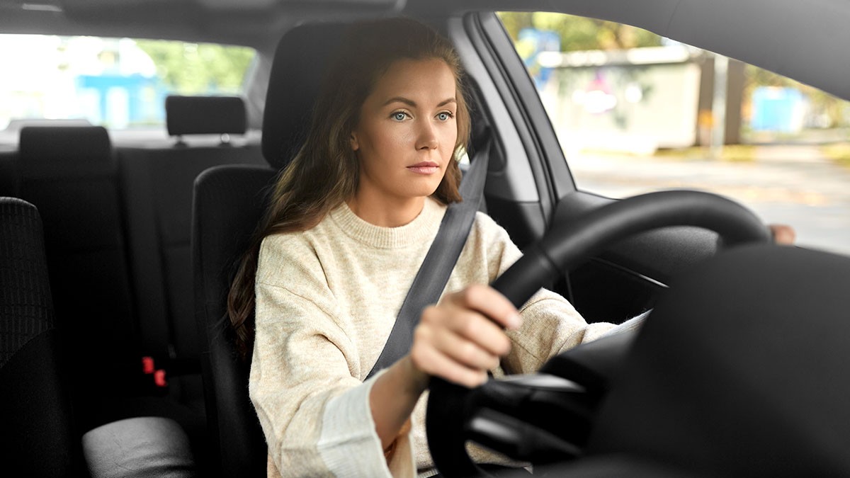 Woman with long hair driving a car, focused on the road. She wears a light-colored top and grips the steering wheel, with a seatbelt fastened.