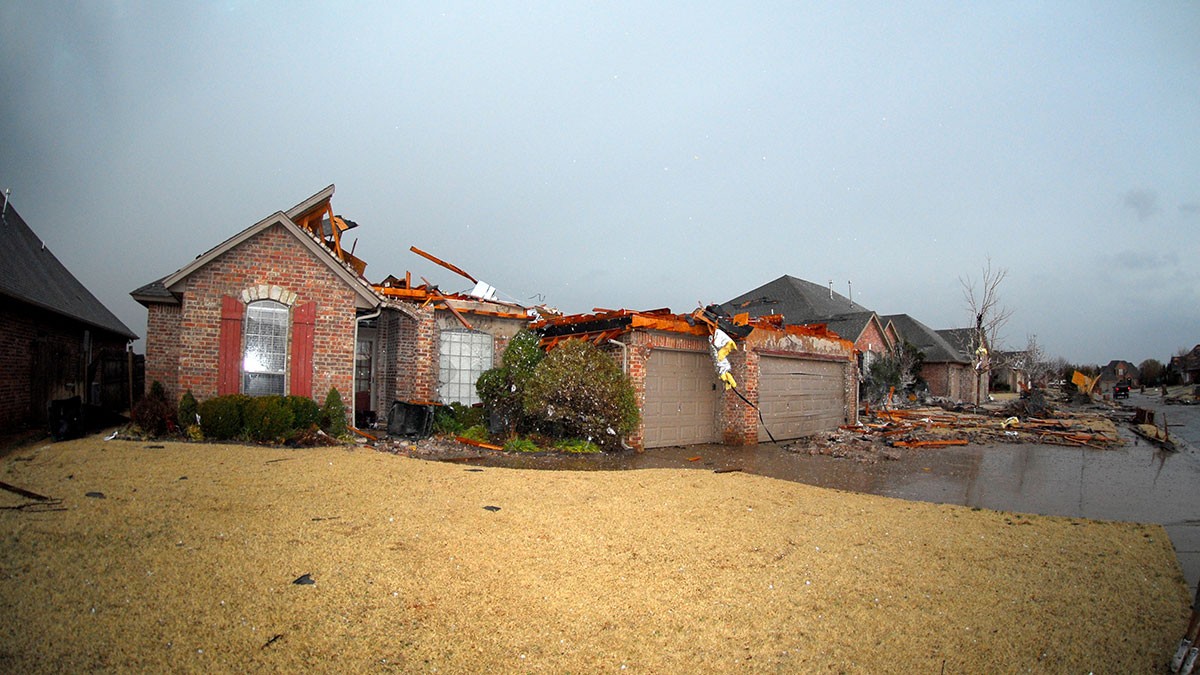 Damaged brick house with a partially destroyed roof, debris scattered, and neighboring houses affected by a storm, under a cloudy sky.