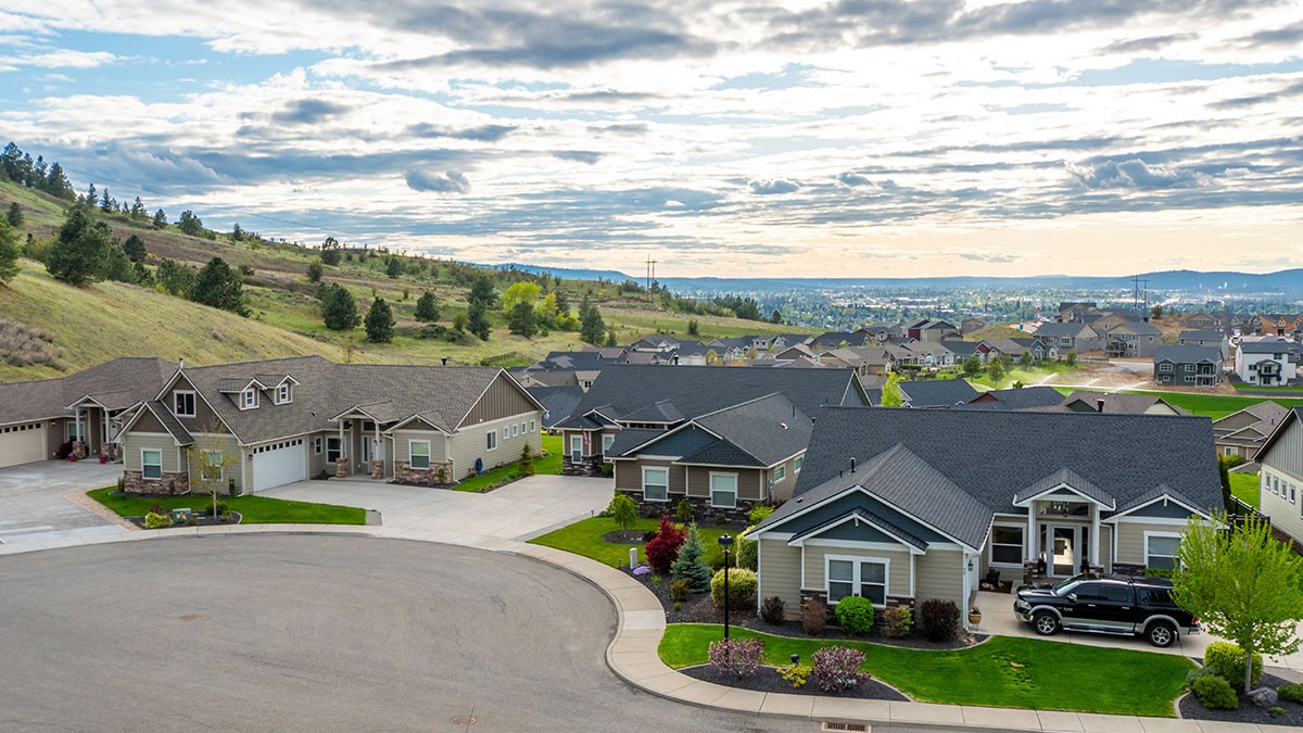 Aerial view of a suburban neighborhood with single-story houses, manicured lawns, and a mountainous backdrop under a cloudy sky.