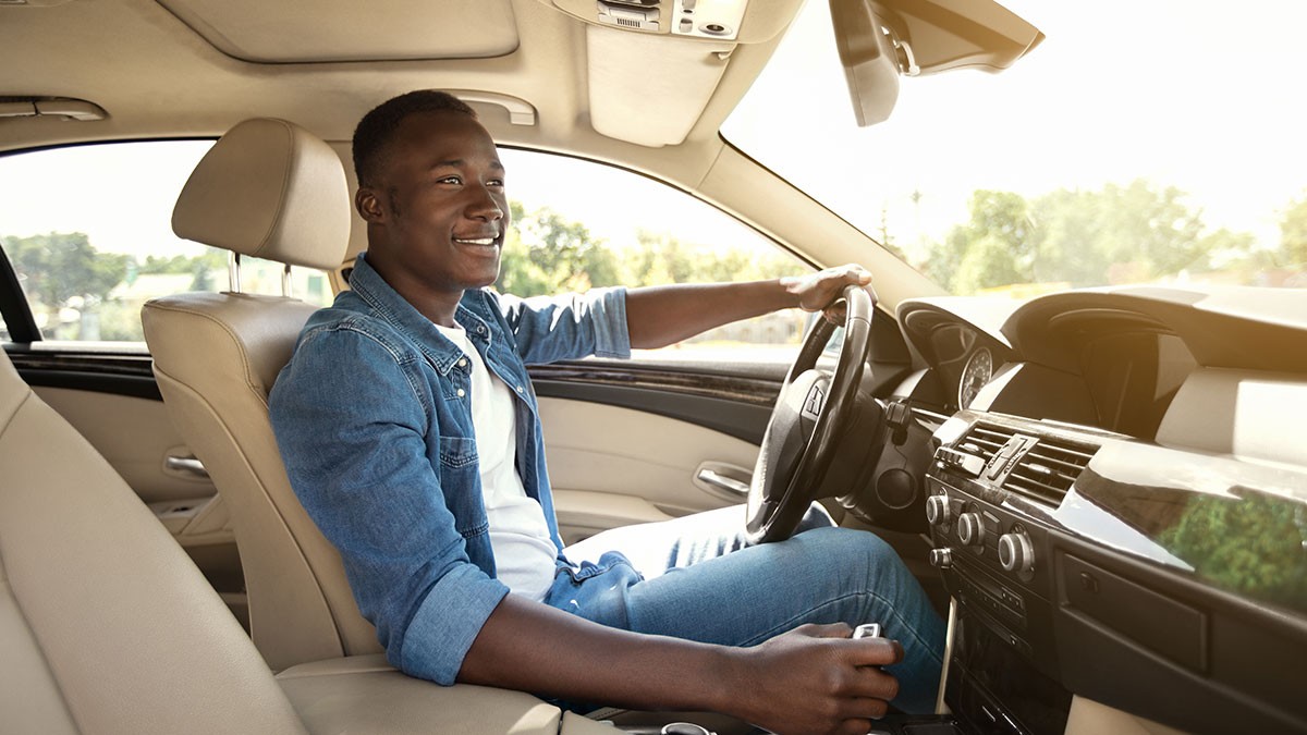 Man in a denim jacket driving a car, smiling, with sunlight streaming through the window. Interior features beige seats and a dashboard.