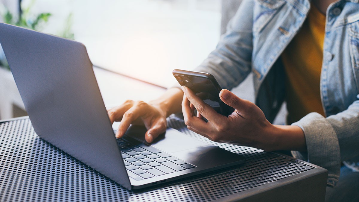 Person using a laptop and holding a smartphone, seated at a perforated metal table, wearing a denim jacket.