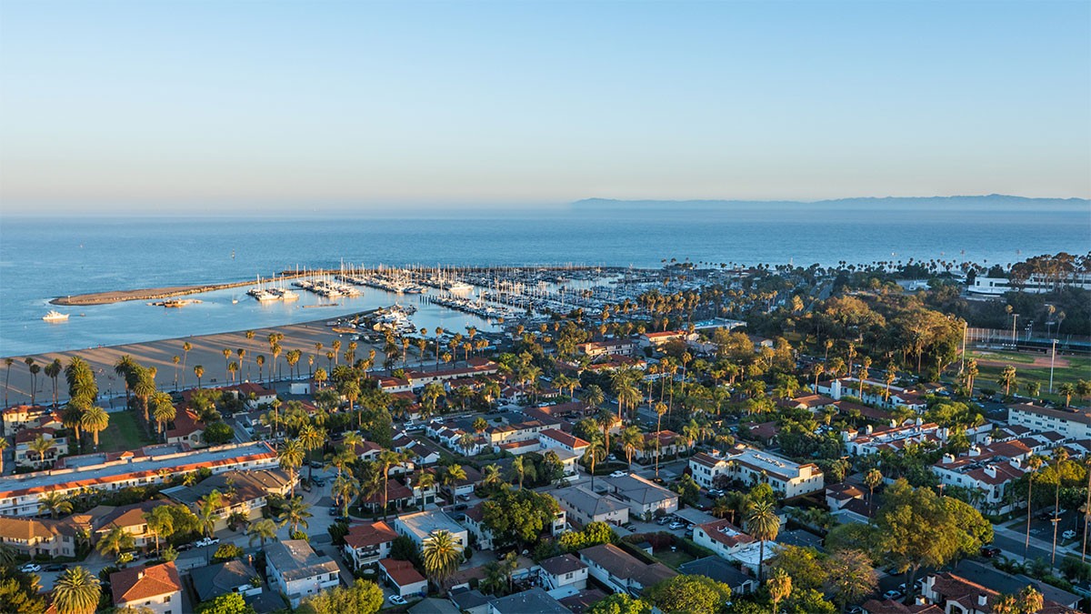Aerial view of a coastal city with a marina, boats, and residential areas surrounded by greenery, under a clear blue sky.