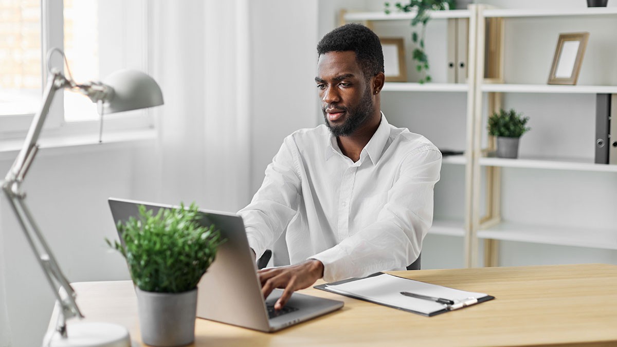 Man in a white shirt working on a laptop at a wooden desk, with a lamp, plants, and shelves in the background.