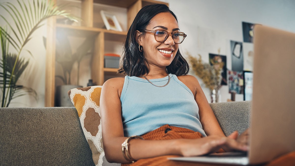 A woman wearing glasses and a blue tank top sits on a couch smiling at her laptop in a cozy living room setting.