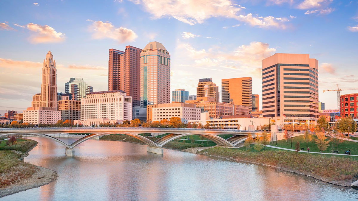 Skyline of a city with modern skyscrapers and a bridge over a river at sunset, reflecting warm colors in the water and sky.