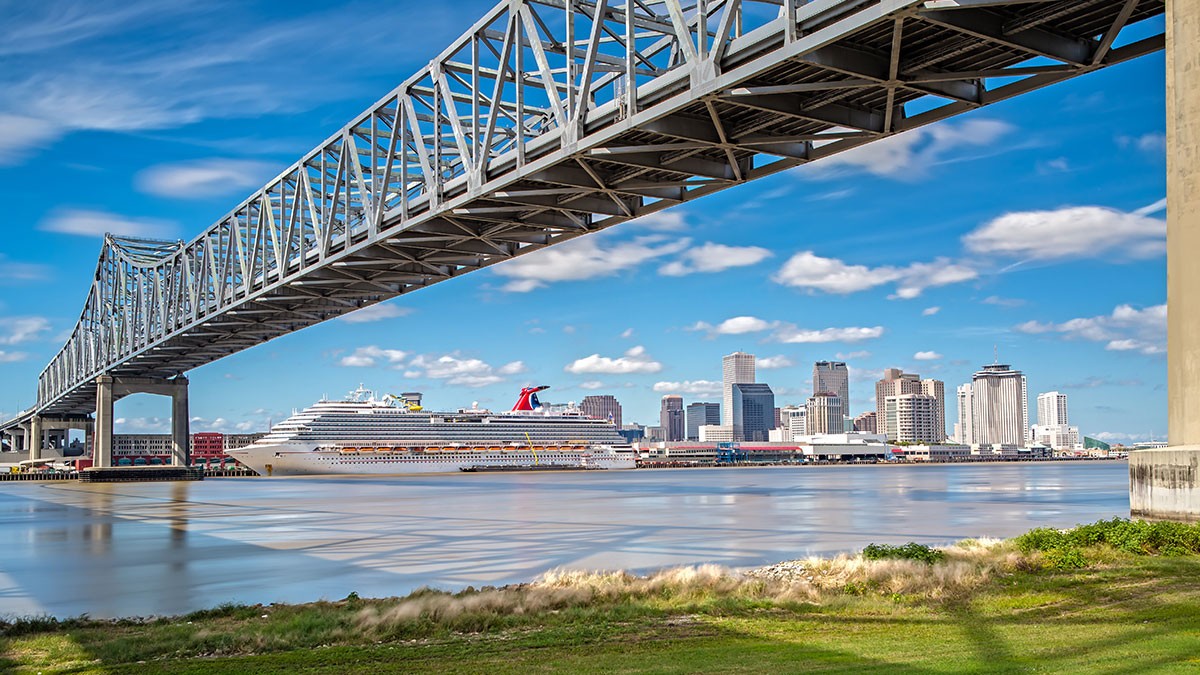 A large cruise ship docked near a city skyline under a steel bridge, with a river in the foreground and a blue sky above.
