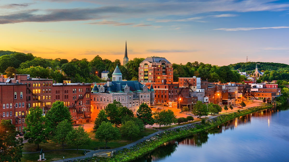 Scenic view of a riverside town at sunset with brick buildings, a church spire, trees, and a reflection on the calm water.