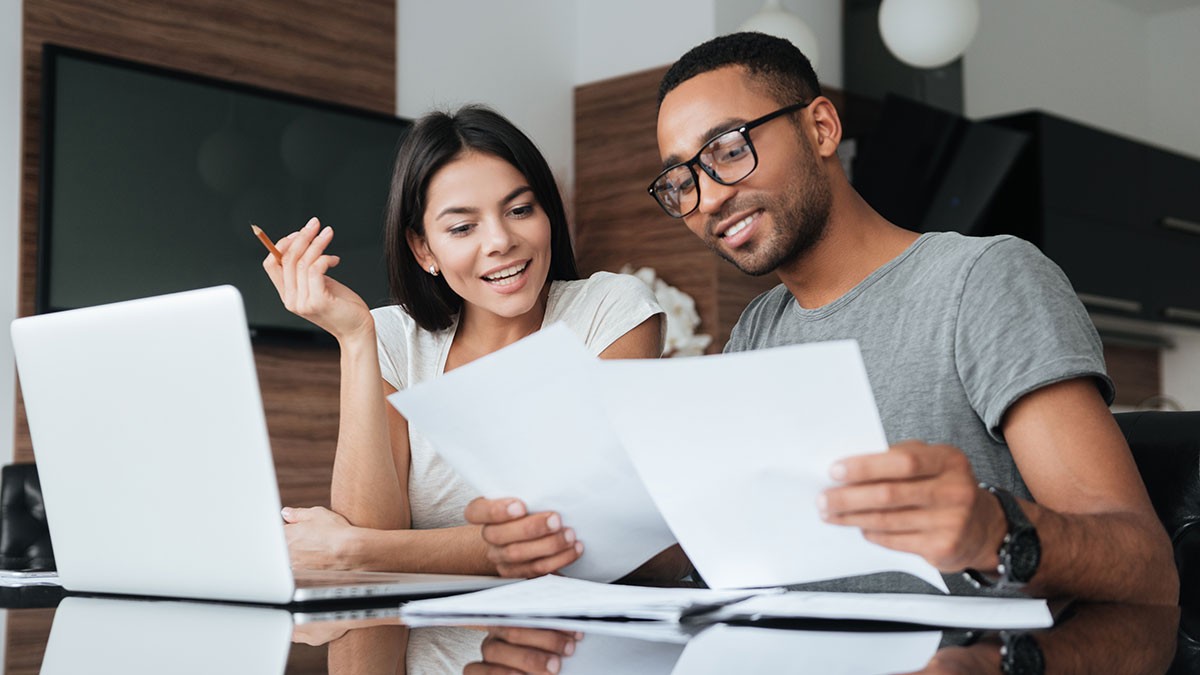 A smiling couple reviews documents together at a table, with a laptop open in front of them in a modern home setting.