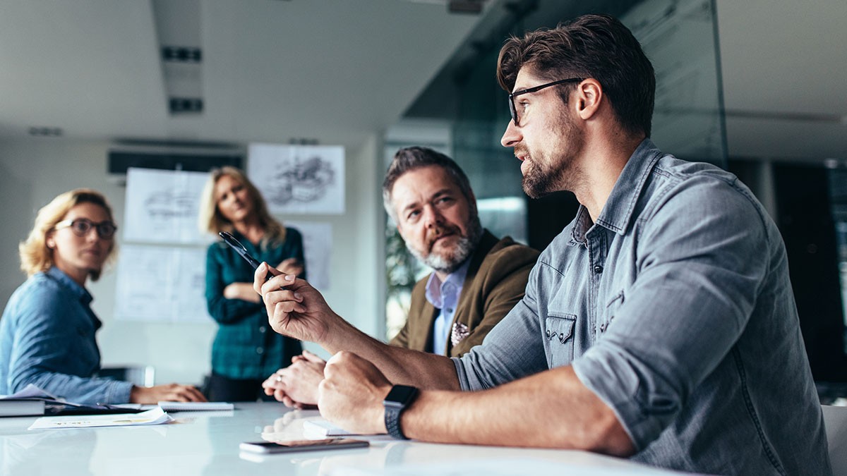 Four colleagues in a meeting: a bearded man with glasses speaks and gestures while three coworkers listen around a conference table.