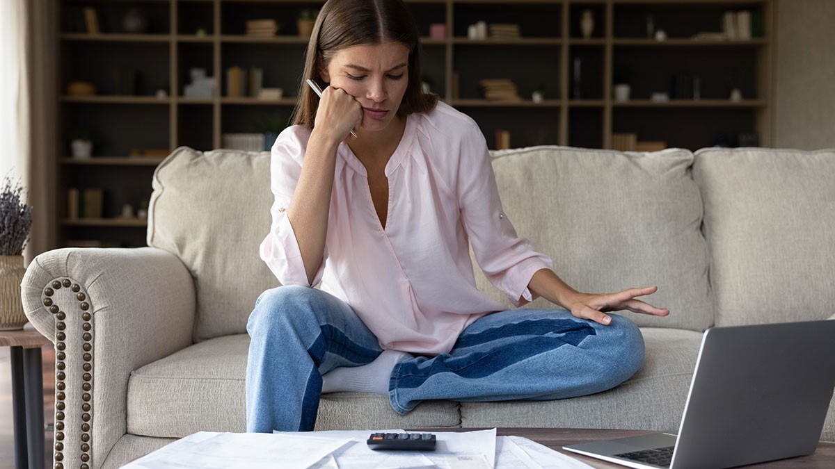 Woman sitting on a couch, looking concerned at a laptop. Papers and a calculator are on the table, suggesting financial or work-related stress.