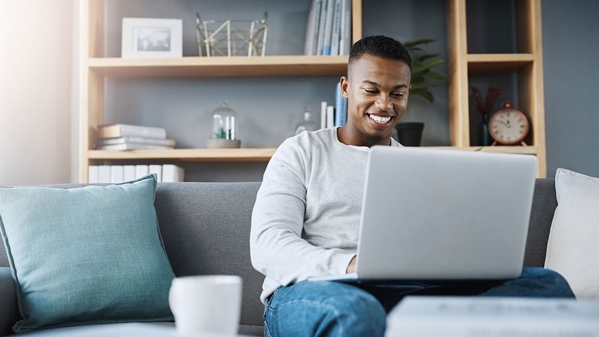 A man sitting on a couch is smiling while using a laptop. Books and a clock are on the shelves in the background.