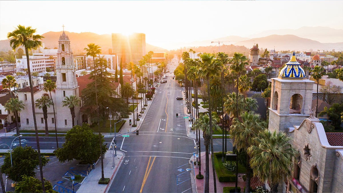 Aerial view of a city street lined with palm trees at sunset, featuring historic buildings and mountains in the background.