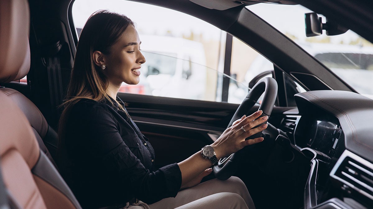 Woman in black top smiling while sitting in the driver's seat of a car, hands on the steering wheel, looking at the dashboard.