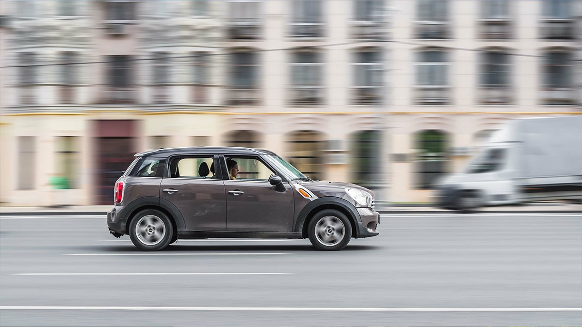 A black Mini Cooper driving on a city street with blurred buildings and a van in the background, indicating motion.