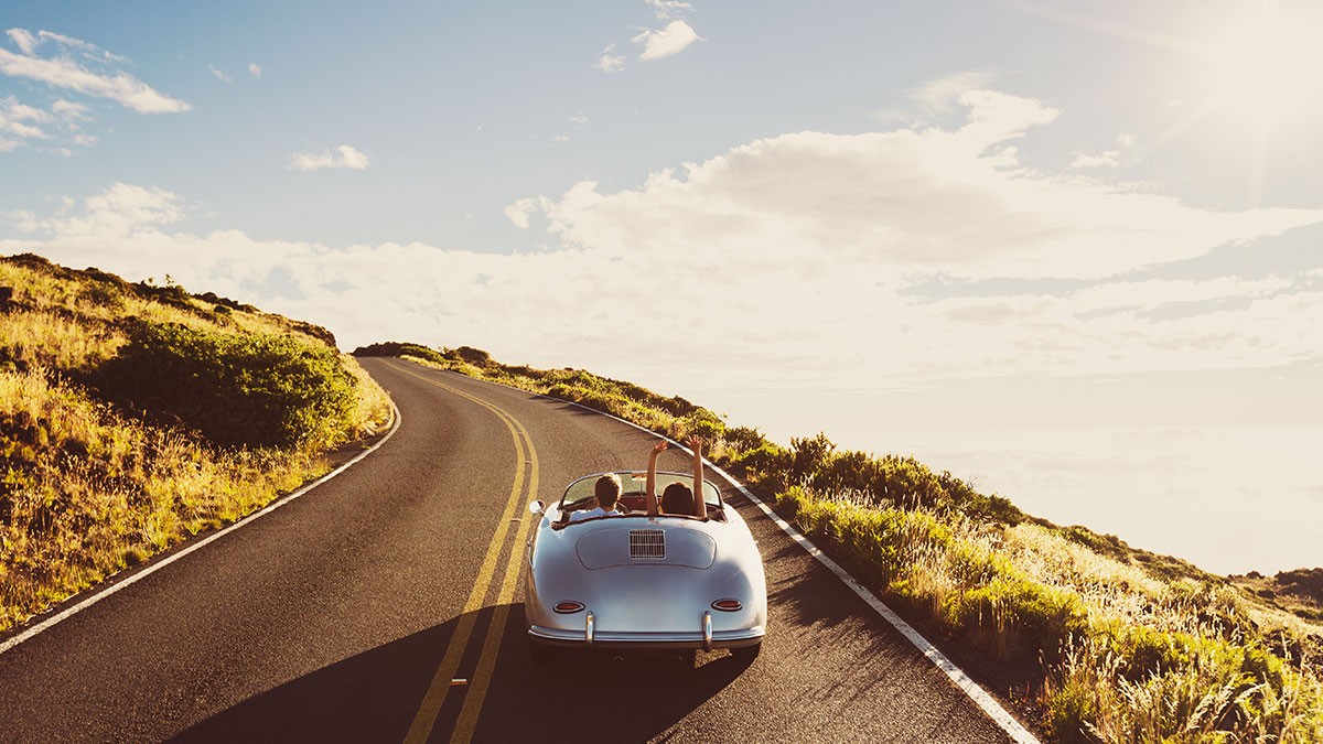 A classic convertible drives along a winding road through scenic hills under a blue sky with scattered clouds.