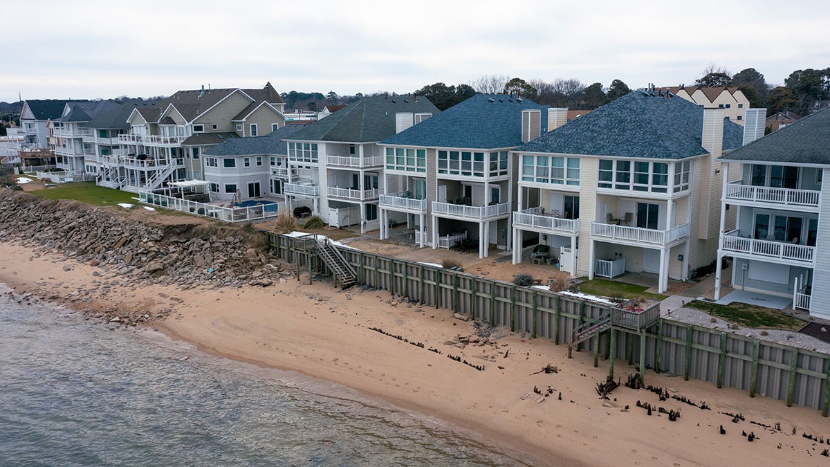 Several beachfront houses with large balconies face a sandy shore, backed by a retaining wall and rocky embankment, under a cloudy sky.