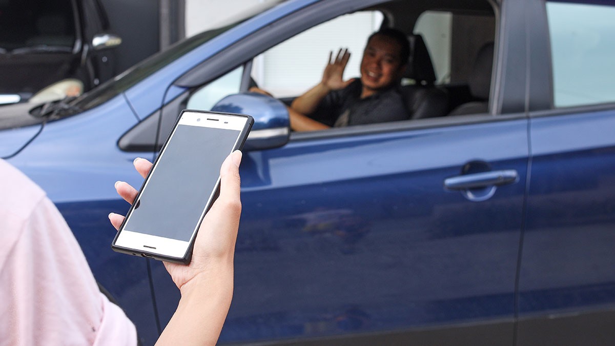 Person holding a smartphone greets a smiling driver in a blue car.