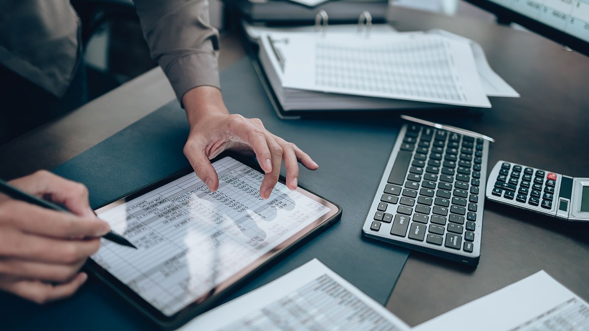Person working with financial documents on a tablet, next to a keyboard and calculator on a desk.