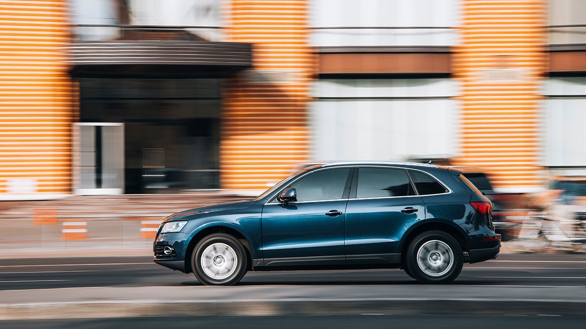 A blue SUV driving quickly on a city street with a blurred background of orange-striped buildings.