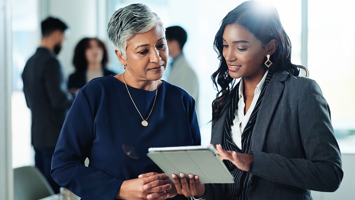 Two women in business attire discuss something on a tablet in an office setting, with colleagues in the background.