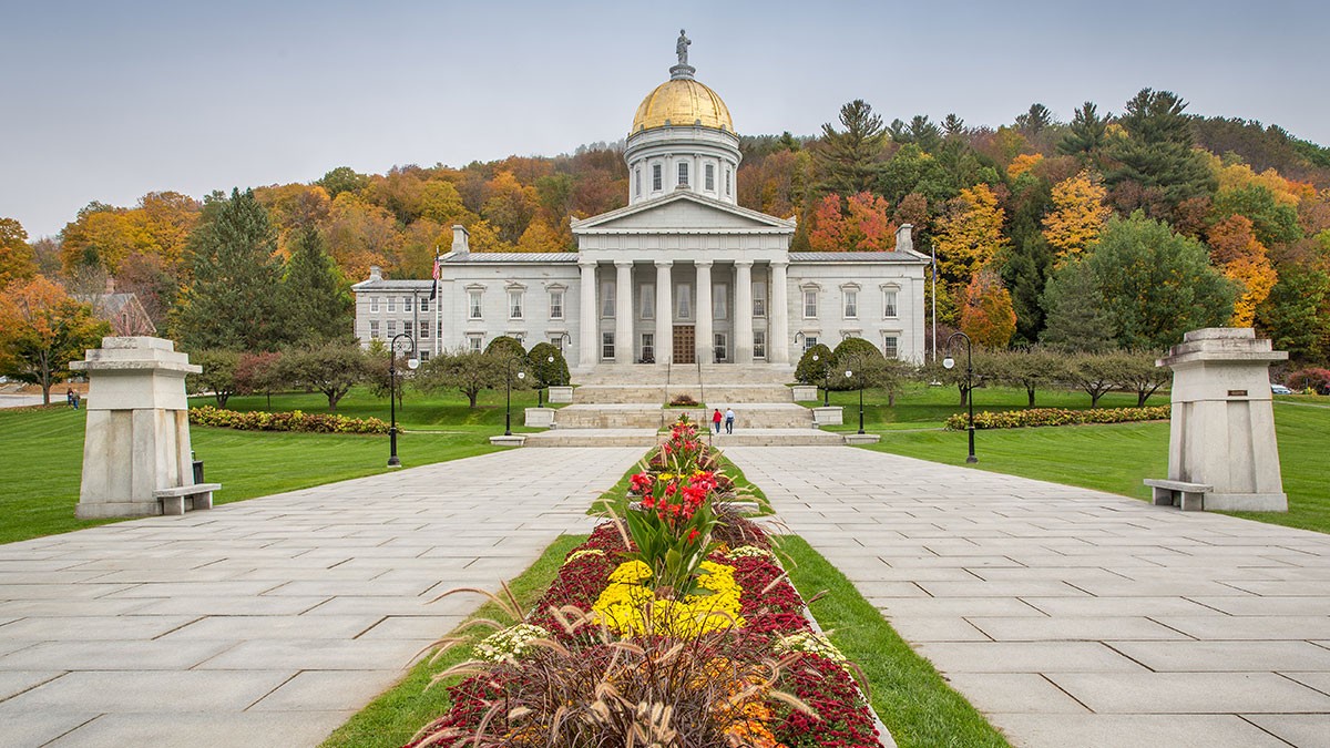 Vermont State House with a gold dome, surrounded by vibrant autumn trees and a landscaped garden path in the foreground.
