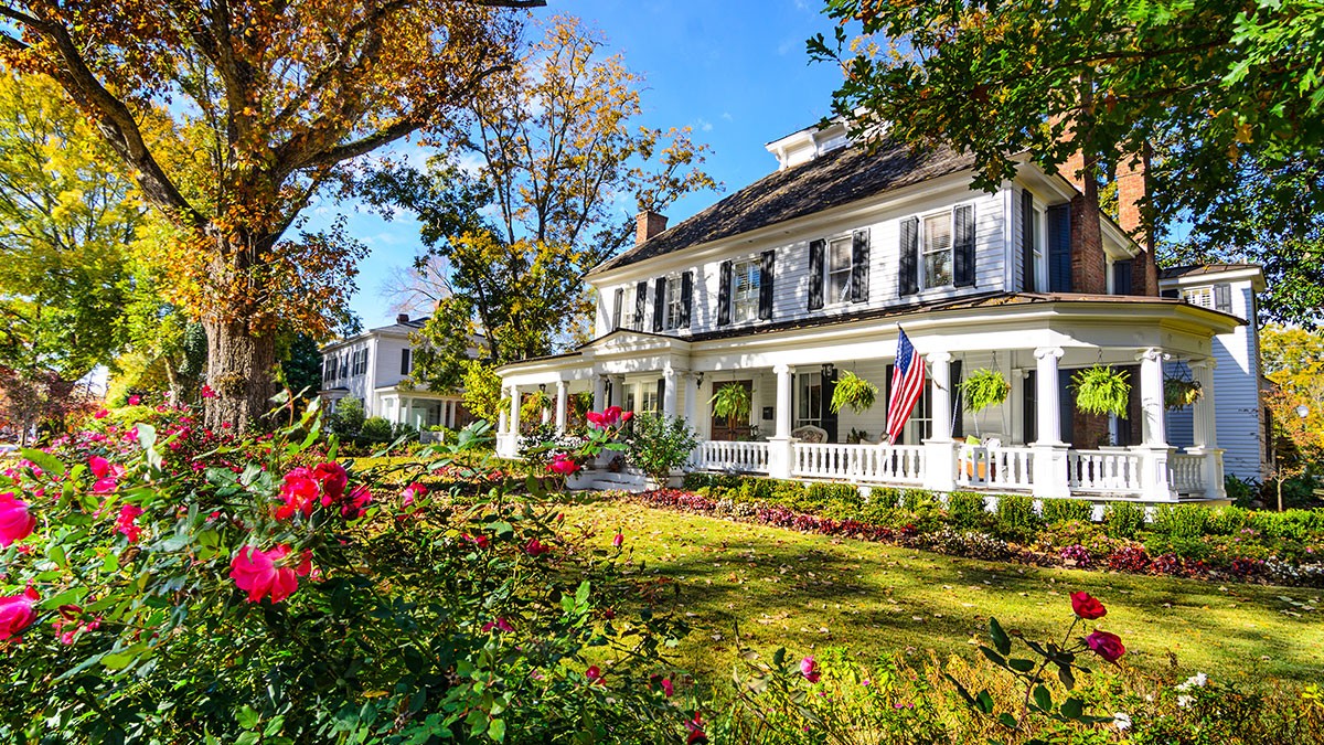 Charming colonial-style house with a wide porch, American flag, lush garden, and autumn trees under a clear blue sky.