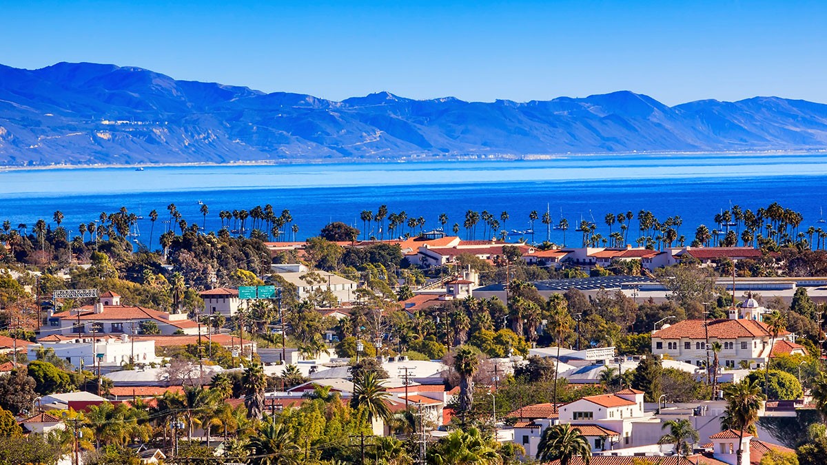 Scenic view of a coastal town with red-roofed buildings, palm trees, and mountains in the background under a clear blue sky.