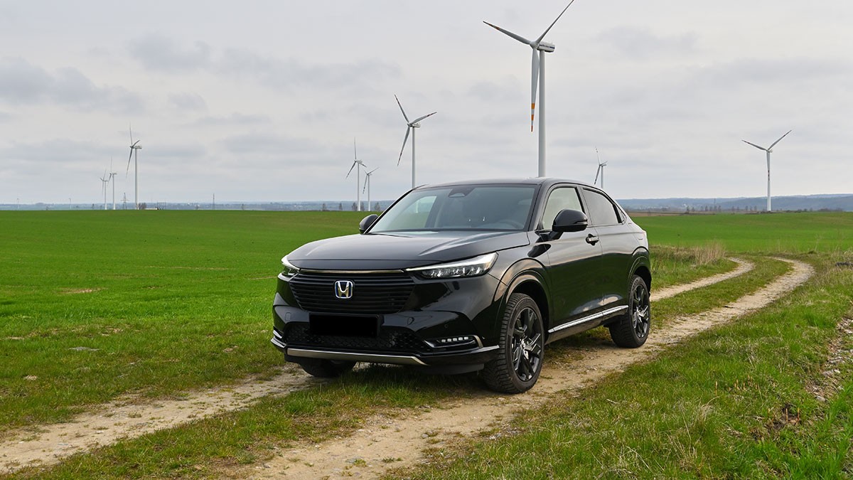 A black SUV parked on a dirt path in a green field with wind turbines in the background under a cloudy sky.